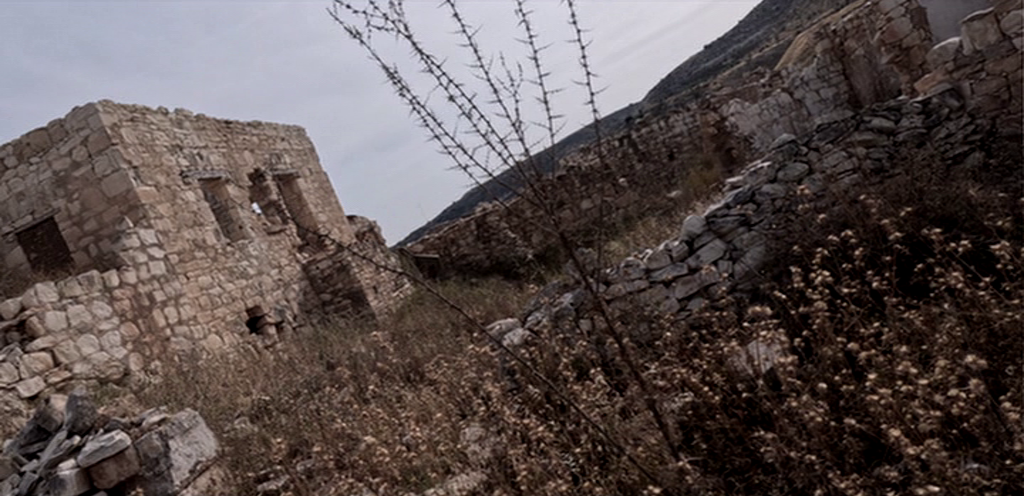 Abandoned stone buildings in a dry, overgrown landscape.